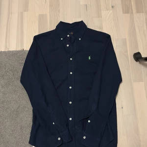 Mörkblå skjorta från Ralph Lauren - Snygg mörkblå skjorta från Ralph Lauren i slim fit-modell. Skjortan har klassisk krage, vita knappar och det ikoniska gröna broderade logotypen på bröstet. Perfekt för dig som gillar stilrena och tidlösa plagg.