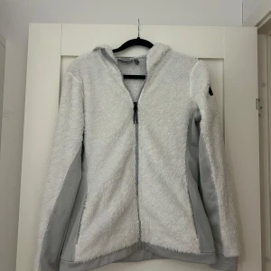 Vit teddyjacka från McKinley med huva - Supermjuk och fluffig vit teddyjacka från McKinley med grå detaljer och hel dragkedja. Jackan har huva och långa ärmar, perfekt för kyliga dagar. Snygg och bekväm att slänga på sig när du vill hålla dig varm och ändå se chill ut. Det grå på teddyjackan känns som ett sportmaterial och är mycket luftigt, denna tröjan är perfekt framför fredagsmyset samt i skidbacken.🍿⛷️