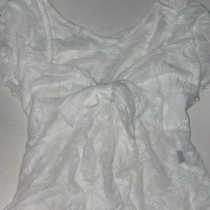 Vit blus med stor rosett - Supersöt vit blus i transparent tyg med en stor rosett framtill. Blusen har korta ärmar och en lätt skir struktur som ger en romantisk vibe. Perfekt för dig som gillar detaljer och vill sticka ut lite extra.