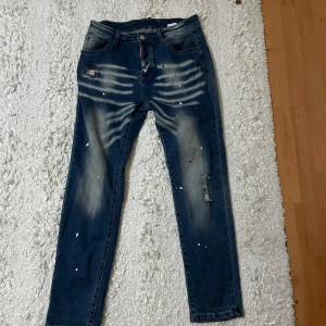 DSQUARED2 blå jeans med färgstänk - Snygga blå jeans från DSQUARED2 med coola färgstänk i vitt, rött och svart samt slitningar och broderad detalj på bakfickan. Jeansen har en rak passform och klassisk femficksdesign. Märkespatch i svart och rött bak på linningen.