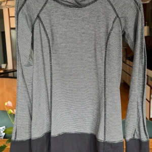 Randig hoodie från Lululemon - Snygg hoodie från Lululemon i svart och grått med tunna ränder. Tröjan har huva, lång ärm och en praktisk ficka med dragkedja på sidan. Materialet är mjukt och stretchigt, perfekt för träning eller chill. Diskret logga på baksidan och kontrastfärg vid muddar och nederkant.