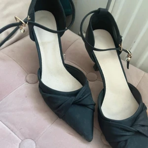 Svarta pumps med smyckesdetalj - Stilrena svarta pumps med spetsig tå och låg klack. Skorna har en elegant knutdetalj framtill och smal ankelrem med guldfärgat spänne och hängande smycke. Perfekta för dig som gillar klassisk och chic stil.