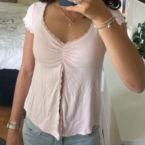 Ljusrosa topp med spets från Gina Tricot - Supersöt ljusrosa topp från Gina Tricot med v-ringning, spetskant och rynkdetalj framtill. Korta ärmar med volangkant och en liten slits framtill. 