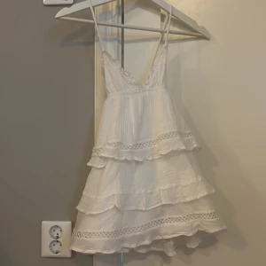 Vit volangklänning från Gina Tricot - Supersöt vit kortklänning från Gina Tricot med tunna axelband och omlott-v-ringning. Klänningen har flera lager med volanger och spetsdetaljer, vilket ger en romantisk och boho känsla. Perfekt för sommarens alla tillfällen! Klänningen är ganska kort så det behövs nog att man har shorts under. Använd fåtal gånger💕