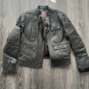 Svart skinnjacka från Jofré - Snygg svart skinnjacka från Jofré med coola detaljer som dragkedjor på ärmarna, flera fickor med dragkedja och tryckknappar samt quiltad axelparti. Jackan har en klassisk bikerstil och är helfodrad med slät insida.