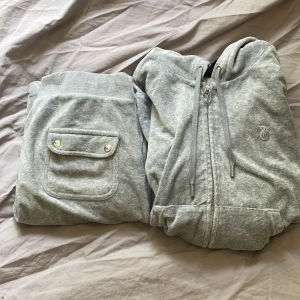 Grå Juicy Couture hoodie med dragkedja - Supermjuk grå hoodie från Juicy Couture med dragkedja, huva och snörning. Klassisk logga broderad på bröstet och snygga metalldetaljer. Perfekt för en chill och trendig look. Materialet är mjukt och skönt, troligtvis bomullsmix. Byxor ingår med 