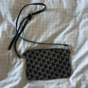 Svart och grå axelväska från Carin Wester - Snygg axelväska från Carin Wester i svart och grått med monogrammönster över hela väskan. Väskan har en smal justerbar axelrem i svart skinn och dragkedja upptill. Insidan är svart med fack för kort och småsaker. Perfekt storlek för det viktigaste.