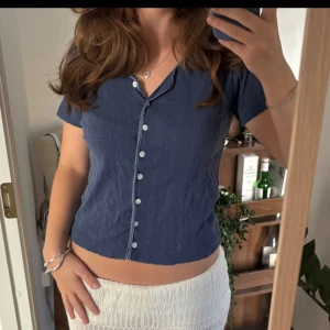 Blå ribbad topp - Snygg blå ribbad topp från Disson med korta ärmar och v-ringning. Dekorativa små knappar längs hela framsidan och virkad kant runt halsen ger en unik touch. 