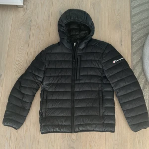 Svart dunjacka från Champion - Snygg svart dunjacka från Champion med huva och dragkedja framtill. Jackan har quiltad design, lång ärm och logga på ena ärmen. Perfekt för kyliga dagar och enkel att matcha med olika outfits. Köpte den för 1000kr och pris kan diskuteras! Använd i 2 månader innan den blev för liten för jag är 190cm