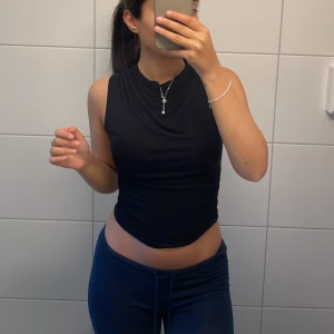 Svart ärmlös topp med drapering - Säljer en svart ärmlös topp med snygg drapering vid halsen. Toppen har en tight passform och är tillverkad i ett mjukt, stretchigt material som sitter skönt mot kroppen. Perfekt att styla med jeans eller kjol för en clean look.