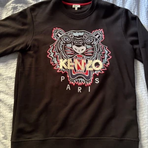Svart sweatshirt med tiger från Kenzo - Kenzo sweatshirt i svart med ikonisk broderad tiger och logga på bröstet. Tröjan har rund hals, långa ärmar och detaljer i rött, grått och beige. Perfekt för dig som vill sticka ut med en fet design och streetstyle-vibe. såklart ÄKTA