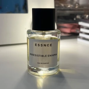 ESSNCE Irresistible Enigma parfym - En stilren parfymflaska från ESSNCE med doften Irresistible Enigma. Flaskan är cylindrisk i genomskinligt glas med svart lock och vit etikett. Innehållet är ljusgult och parfymen är en Eau de Parfum. Perfekt för dig som gillar minimalistisk design.