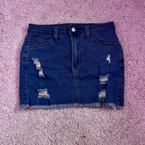 Mörkblå jeanskjol med slitningar - Snygg mörkblå jeanskjol med fransad nederkant och slitna detaljer framtill. Kjolen har klassiska fickor både fram och bak samt hällor för bälte. Plus stretchigt material.