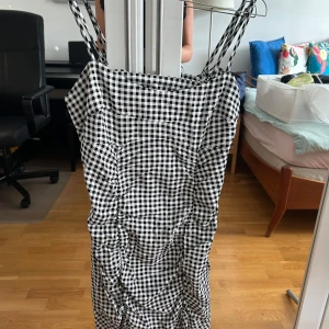 Svartvit rutig klänning från H&M Divided - Säljer en svartvit rutig kortklänning från H&M Divided i storlek 40. Klänningen har smala axelband och snygga rynkade detaljer framtill. Perfekt för dig som gillar en trendig och lekfull stil med klassisk rutigt mönster.