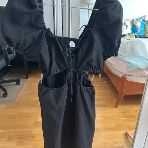 Svart kortklänning med puffärm - Svart kortklänning från H&M i storlek 40. Klänningen har puffiga kortärmar, smock upptill och snygga cut-outs vid midjan. Materialet är lätt och har en lite skrynklig struktur. Knytning framtill ger en extra detalj. Perfekt för dig som gillar trendiga och unika plagg.