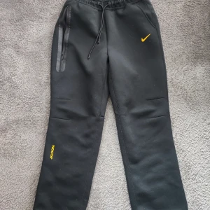 Nike NOCTA svarta mjukisbyxor M - Svarta mjukisbyxor från Nike NOCTA med gul logga och detaljer. Byxorna har dragsko i midjan, dragkedjeficka och broderade NOCTA- och Nike-symboler. Sköna och stilrena, perfekta för chill eller streetwear.