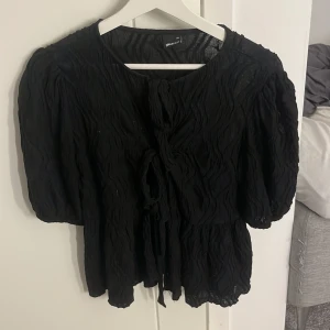 Svart blus med knyt från Gina Tricot - Säljer en svart blus från Gina Tricot med puffiga ärmar och snygg knytning framtill. Blusen har en vågig, strukturerad yta och är lätt och luftig. Perfekt för dig som gillar trendiga detaljer och en cool siluett.