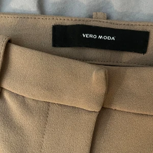Beige kostymbyxor från Vero Moda - Stilrena beige kostymbyxor från Vero Moda med raka ben och klassiska hällor för bälte. Byxorna har en snygg midja och är perfekta för en clean och dressad look. Materialet känns mjukt och har en lätt struktur.