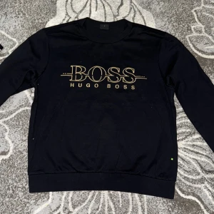 Svart tröja från Hugo Boss - Säljer en stilren svart tröja från Hugo Boss med guldigt tryck på framsidan. Tröjan har en slim fit passform och långa ärmar. Perfekt för en snygg och avslappnad look.