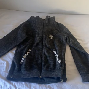 Svart zipjacka från Philipp Plein - Svart zipjacka från Philipp Plein med vita logotexter på fickorna och emblem på bröstet. Jackan har huva, hel dragkedja och ribbade muddar. Perfekt för dig som gillar streetwear och vill sticka ut med snygga detaljer.