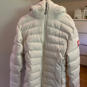 Vit pufferjacka från Huski Wear - Säljer en snygg vit pufferjacka från Huski Wear med huva och dragkedja framtill. Jackan har quiltad design och tydlig logga på ärmen. 