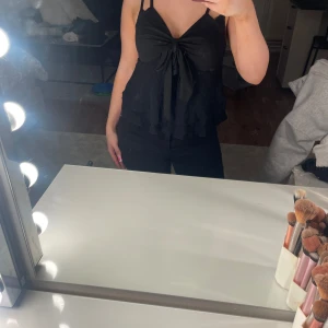 Svart linnetopp med rosett och volang - Supersöt svart linnetopp med smala axelband, stor rosett framtill och dubbla volanglager nedtill. Toppen har v-ringning och är figurnära upptill, perfekt för att skapa en trendig look. Passar dig som gillar feminina detaljer och stilren design.