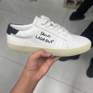 Saint Laurent vita sneakers i skinn - Snygga vita sneakers från Saint Laurent i skinn med svart detalj på hälen och handskriven logga på sidan. Klassisk låg modell med snörning och platt sula i beige ton. Perfekta för dig som gillar stilrena och exklusiva skor med en twist. Strl 44 men passar också 43.