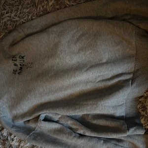 Grå hoodie med tryck från Gina Tricot i storlek 158/164 - Säljer en grå hoodie från Gina Tricot med svart tryck 'JE M'EN AMOUR' och en stjärna på ryggen. Tröjan har huva, ribbade muddar och är tillverkad i mjuk bomullsmix. Perfekt för chill dagar och streetstyle. 