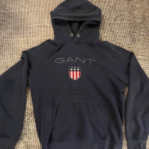 Marinblå hoodie från GANT med emblem - Snygg marinblå hoodie från GANT med stort broderat emblem och logga på bröstet. Tröjan har huva med snörning, känguruficka och långa ärmar. Materialet är mjuk bomull och polyester, perfekt för chill dagar. Klassisk amerikansk stil med coola detaljer.