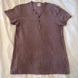 Lila t-shirt med v-ringning och knappar - Snygg lila t-shirt med v-ringad hals och tre svarta knappar framtill. T-shirten har korta ärmar och är gjord i mjuk bomull, perfekt för en chill och avslappnad stil. Passar dig som gillar enkel design med en liten twist.