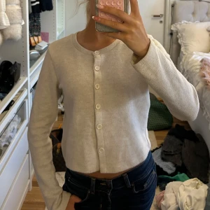 Beige kofta från Zara med knappar - Säljer en beige, croppad kofta från Zara med rund hals och långa ärmar. Koftan har knappar hela vägen framtill och är gjord i mjukt stickat smått ribbat material som känns skönt mot huden. Perfekt att slänga över en topp för en chill och clean look.