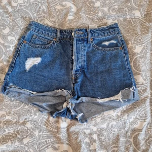 Blå jeansshorts med slitningar H&M - Säljer ett par blå jeansshorts från H&M i storlek 38. De har hög midja, klassiska fem fickor och coola slitningar framtill och baktill. Rå kant nedtill ger en avslappnad vibe. Perfekta för varma dagar och festivalhäng.