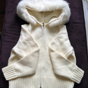 Fur jacket  - Mysig vit stickad jacka med dragkedja och stor huva kantad med fluffig fuskpäls. Jackan har ribbade muddar och nederkant, perfekt för kalla dagar. Enkel och stilren design som ger en soft vibe.