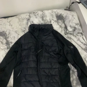 Svart pufferjacka från McKinley - Svart pufferjacka från McKinley med quiltad framsida och dragkedja. Jackan har hög krage och långa ärmar, perfekt för kyliga dagar. Snygg och enkel design som funkar till det mesta. Materialet är syntet och jackan har praktiska fickor med dragkedja.