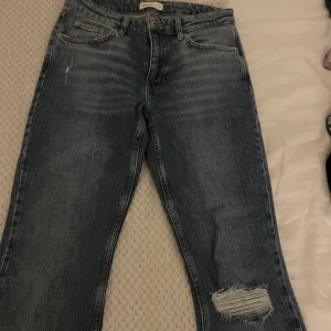 Blå jeans med slitning på knät - Snygga blå jeans med en rejäl slitning på ena knät och klassisk femficksdesign. Jeansen har en rak passform och är tillverkade i denim. Perfekta för en avslappnad och trendig look.
