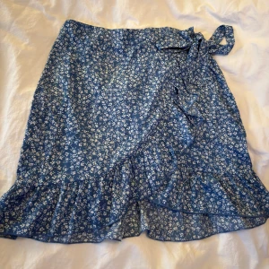 Blå blommig omlottkjol med volang - Supersöt blå kortkjol med vitt blommönster och volangkant. Kjolen har omlottmodell med knyt i midjan som ger en snygg och avslappnad look. Perfekt för vår och sommar!