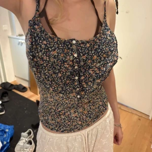 Blommig mörkblå linnetopp med knappar - Supersöt mörkblå linnetopp med små blommor i rosa, orange och beige. Toppen har smala axelband med knyt och en rad vita knappar framtill. Sitter tight och har en feminin v ringad form. Perfekt för varma dagar. Storlek M, använd men sparsamt. 