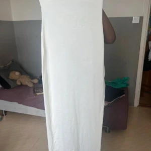 Vit axelbandslös långklänning - Säljer en stilren vit långklänning med axelbandslös design. Klänningen har en slim passform som sitter snyggt mot kroppen och är perfekt för dig som gillar minimalistisk stil. Den har även en slits vid sidan om som en extra detalj