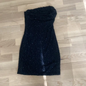 Svart glittrig axelbandslös klänning - Säljer en svart, glittrig kortklänning med axelbandslös design. Klänningen har en tight passform och är täckt av små glittrande detaljer som ger en riktigt snygg look. Perfekt för dig som vill sticka ut och känna dig extra glam. Materialet känns mjukt och följsamt mot kroppen. Jag vet inte vilket varumärke de är för jag har köpt den i albanien