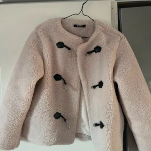 Beige teddyjacka med knäppning - Mysig beige teddyjacka från Gina Tricot med svarta dekorativa knäppningar framtill. Jackan har rund halsringning och är helfodrad, perfekt för kyliga dagar. Långärmad och i en fluffig, trendig stil som passar till många outfits.