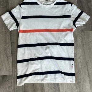 Vit randig t-shirt från Stromberg - Snygg vit t-shirt från Stromberg i storlek S med breda marinblå och en röd rand över bröstet. Klassisk rund hals och korta ärmar. Tillverkad i mjuk bomull som känns skön mot huden. Perfekt för dig som gillar stilrena och fräscha plagg.