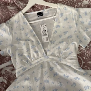 Vit blommig omlottklänning från Gina Tricot - Supersöt kort omlotttopp från Gina Tricot i vit med ljusblått blommigt mönster. Toppen har v-ringad hals, korta ärmar och knyt i midjan för en snygg passform. Perfekt för sommarens alla tillfällen!