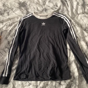 Svart Adidas långärmad tröja XS - Svart långärmad tröja från Adidas med klassiska vita ränder längs ärmarna och vit ribbad krage. Tröjan har Adidas logga på bröstet och är gjord i mjuk bomull. Perfekt för dig som gillar sportig och stilren look.