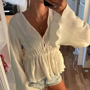 Vit plisserad blus med v-ringning - Supersöt vit blus med plisserat tyg och v-ringning. Vida, långa ärmar med vågig kant och smock i midjan som ger en snygg siluett. Luftig och flowy känsla, perfekt till jeansshorts eller kjol. Ger en romantisk och trendig vibe.