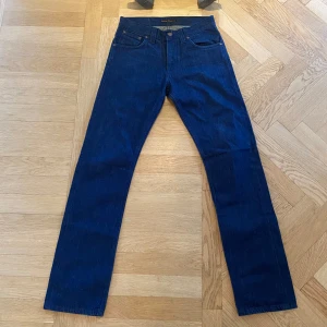 Mörkblå jeans från Nudie Jeans, 30/34 - Snygga mörkblå jeans från Nudie Jeans i klassisk rak modell. Jeansen har fem fickor, orangea detaljer och Nudie-logga på bakfickan samt läderpatch i midjan. Tillverkade i slitstarkt bomullsjeansmaterial. Perfekta för dig som gillar stilrena och tidlösa jeans.