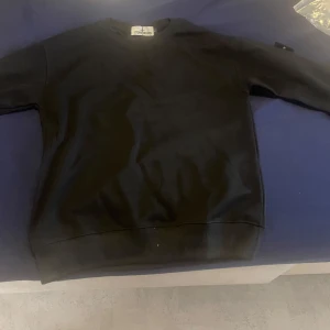 Svart sweatshirt från Stone Island - Svart sweatshirt från Stone Island med klassisk rund hals och ribbade muddar. Tröjan har den ikoniska Stone Island-loggan på vänster ärm. Materialet är mjuk bomull och passformen är normal. Perfekt för dig som gillar stilrena och snygga streetwear-plagg.