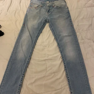 Ljusblå true religion jeans med röd brodyr - Säljer ett par ljusblå jeans med snygg röd brodyr på bakfickorna och klassisk femficksdesign. Jeansen har raka ben och normal passform, tillverkade i mjukt denimtyg. Perfekta för dig som gillar detaljer och vill sticka ut lite extra.