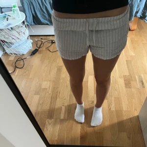Randiga vita shorts med snörning - Säljer ett par vita shorts med tunna grå ränder och snörning i midjan ifrån HM i storlek XS. De har en relaxed fit och är tillverkade i ett lätt och luftigt bomullstyg, perfekta för varma dagar. Shortsen har elastisk midja och är riktigt sköna att bära.