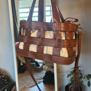 Axelväska  - Säljer en unik axelväska från Marimekko med bruna skinnremmar och beige tyg. Insidan har ett snyggt blommigt mönster i rött och vitt. Väskan har både handtag och axelrem, samt coola gulddetaljer och tryckknappar. Perfekt för dig som vill sticka ut med en stilren men lekfull look. Man kan vända insidan ut och in så de blir blommigt på utsidan💌