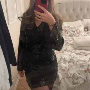 Svart paljettklänning från Gina Tricot - Supersnygg svart kortklänning med blommigt paljettmönster från Gina Tricot. Klänningen har v-ringning, långa transparenta ärmar och sitter tight. Perfekt för dig som vill sticka ut med lite extra glitter och glam. Materialet är polyester och paljetter.